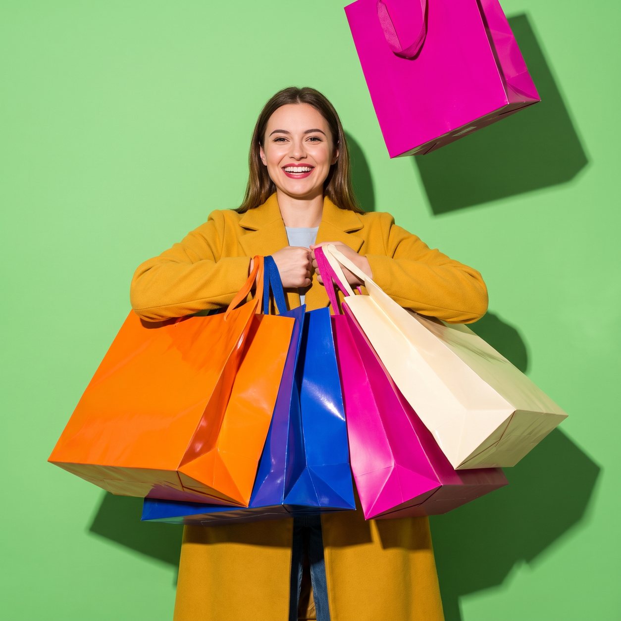 Een lachende vrouw in een mosterdgele jas houdt een armvol glanzende, extra grote tassen in oranje, kobalt, magenta en crème vast, tegen een limoengroene achtergrond.